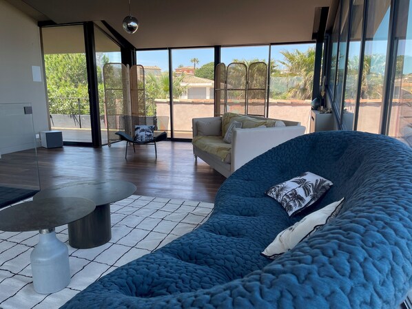 A spacious living area with a large blue sofa, a white couch, and a round table. There is a chair with a patterned cushion, and the room has large windows offering a view of palm trees and buildings.
