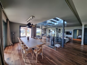 A modern interior with a large wooden dining table, glass walls, and a view of the outdoors.