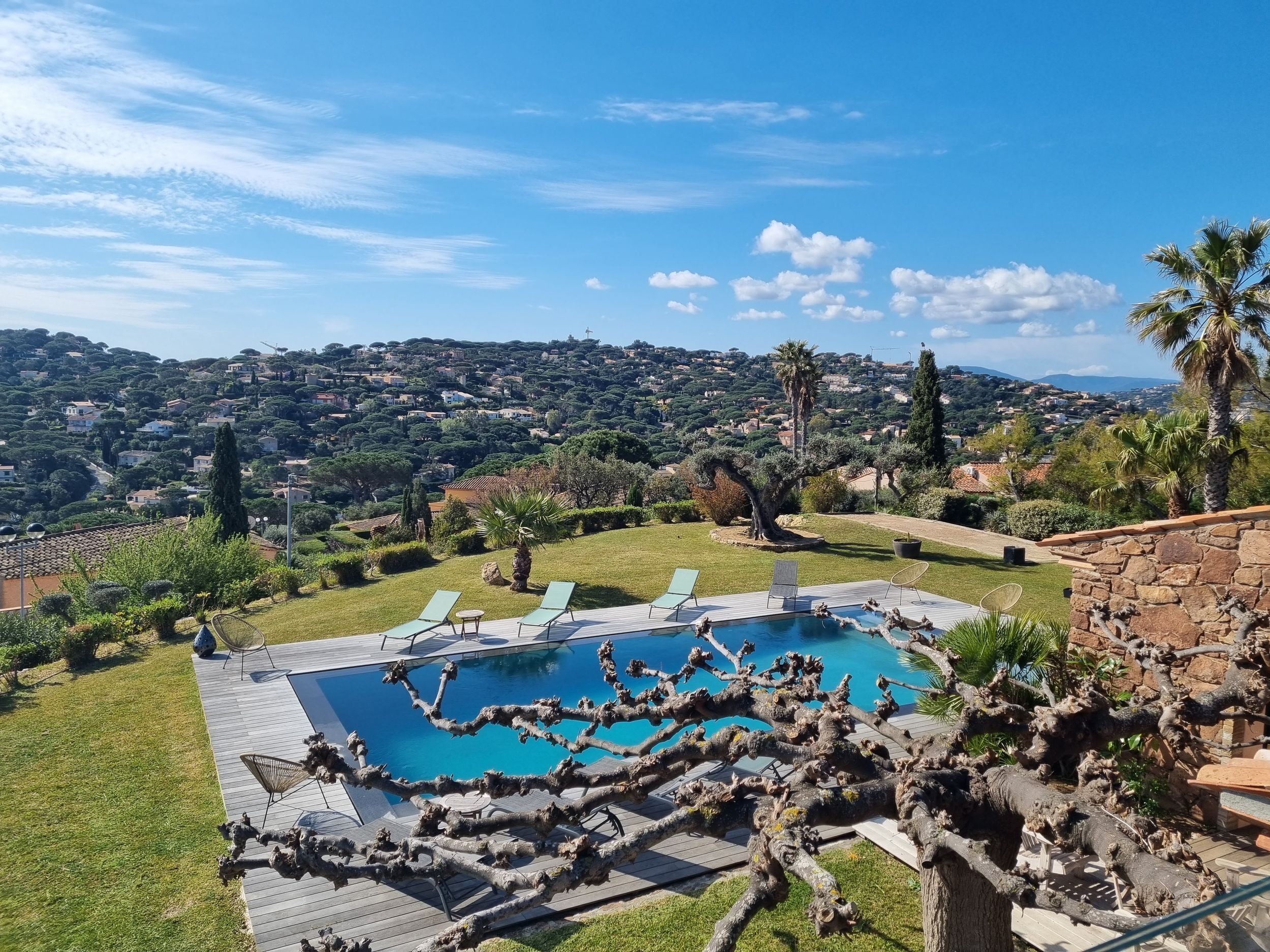 Une piscine entourée de chaises longues, elle-même bordée d’un jardin aménagé et offrant une vue sur une ville perchée sur une colline.
