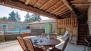 An outdoor patio with a round wooden table and chairs, a pool, stone walls, and a wooden staircase.