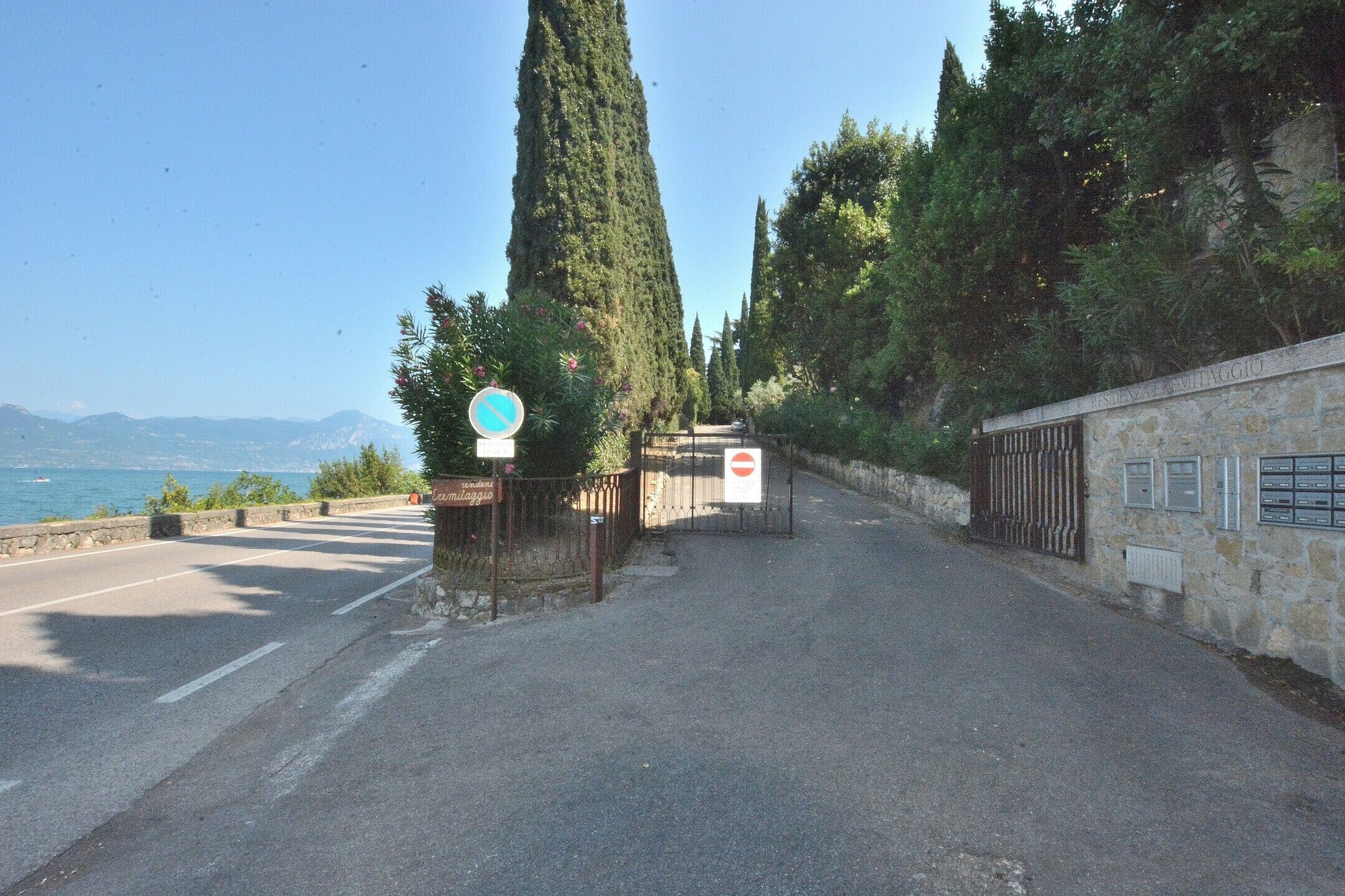 A road leading to a stone building with a gate, surrounded by trees and a clear sky.