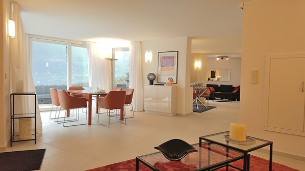 A modern living room with a dining area, a kitchen in the background, and a large window providing natural light.