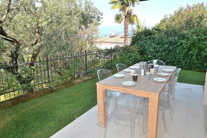 An outdoor dining area with a wooden table and clear chairs, surrounded by a well-maintained garden and a metal fence. - Villa Lisi With Pool (Bardolino)