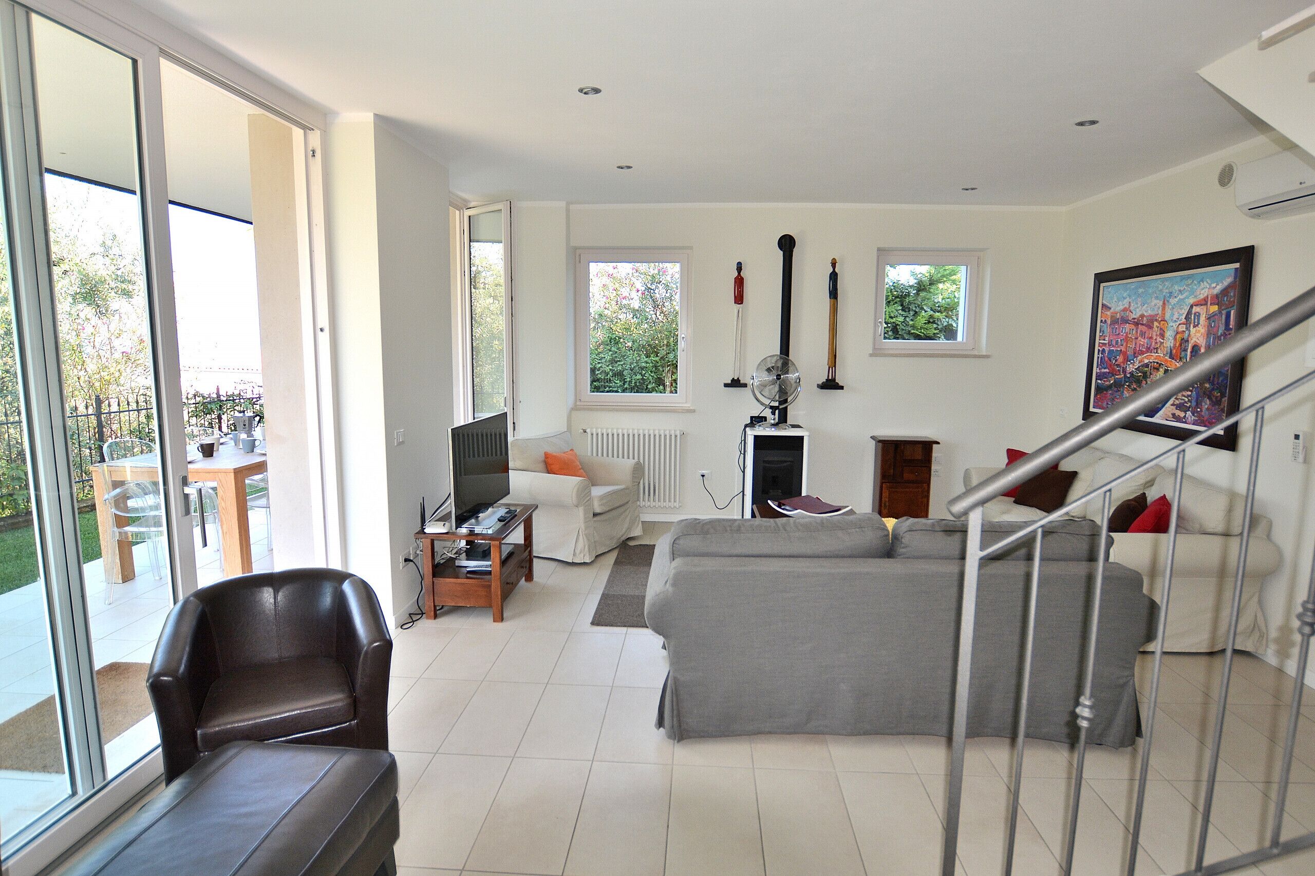 A spacious living room with a grey sofa, a brown leather chair, a wooden coffee table, and a television. There is a staircase with a metal railing leading to an upper level.