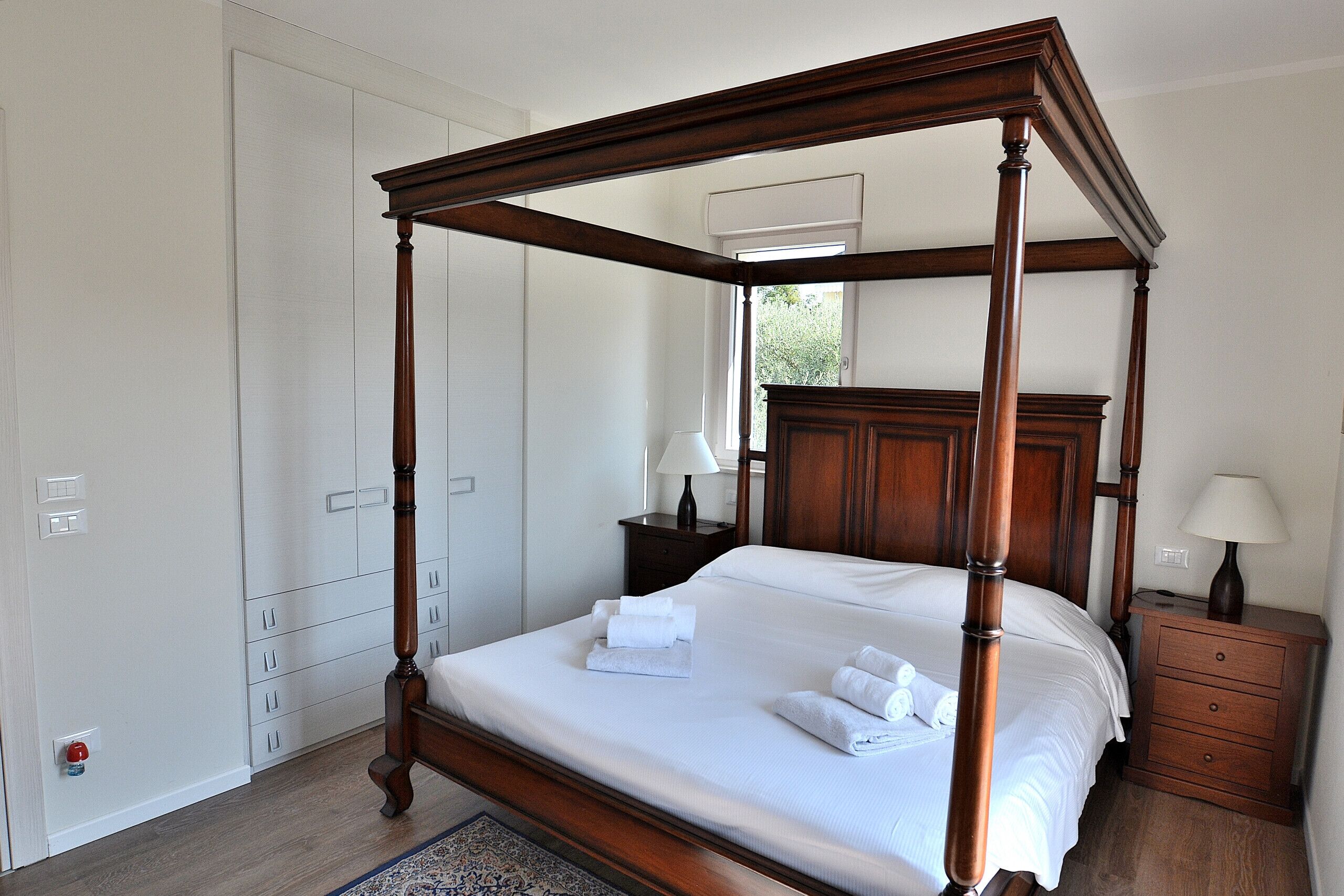 A four-poster bed with white linens, a wooden nightstand with a lamp, and a wardrobe with multiple doors.