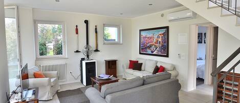 A spacious living room with a grey sofa, white armchairs, and a dining table. There is a staircase leading to another level, and a large painting on the wall.