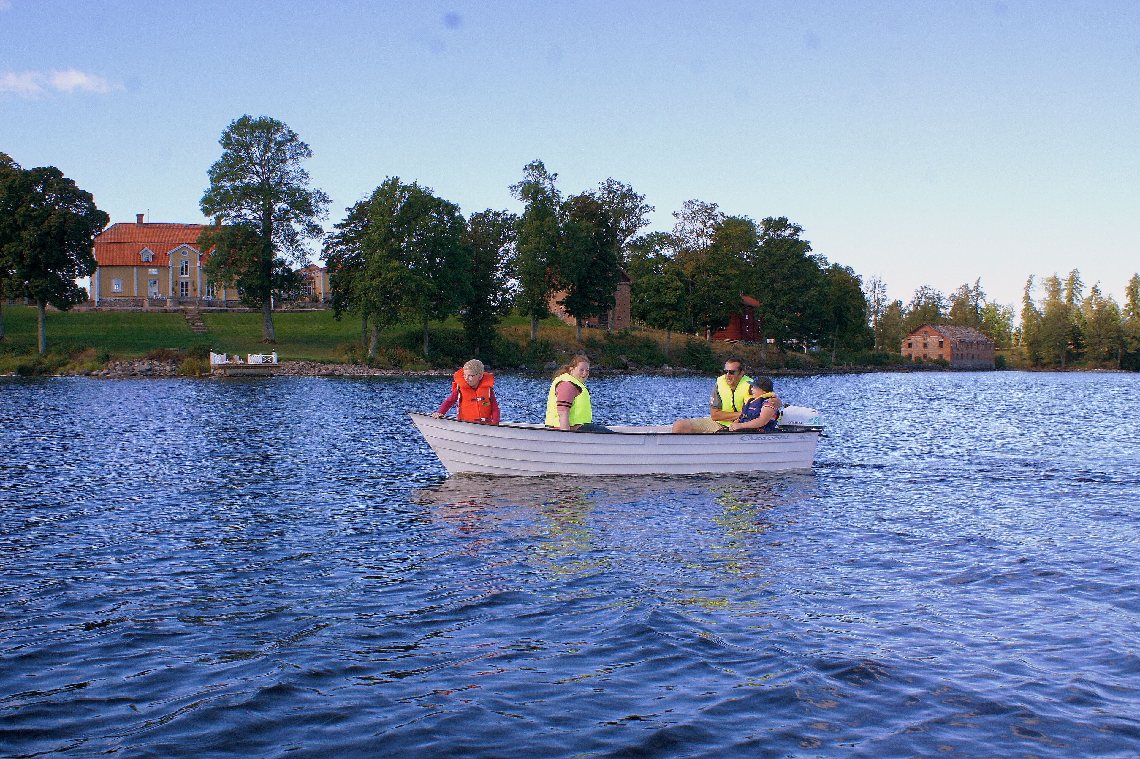 A small boat with three people on a calm lake, surrounded by trees and buildings.