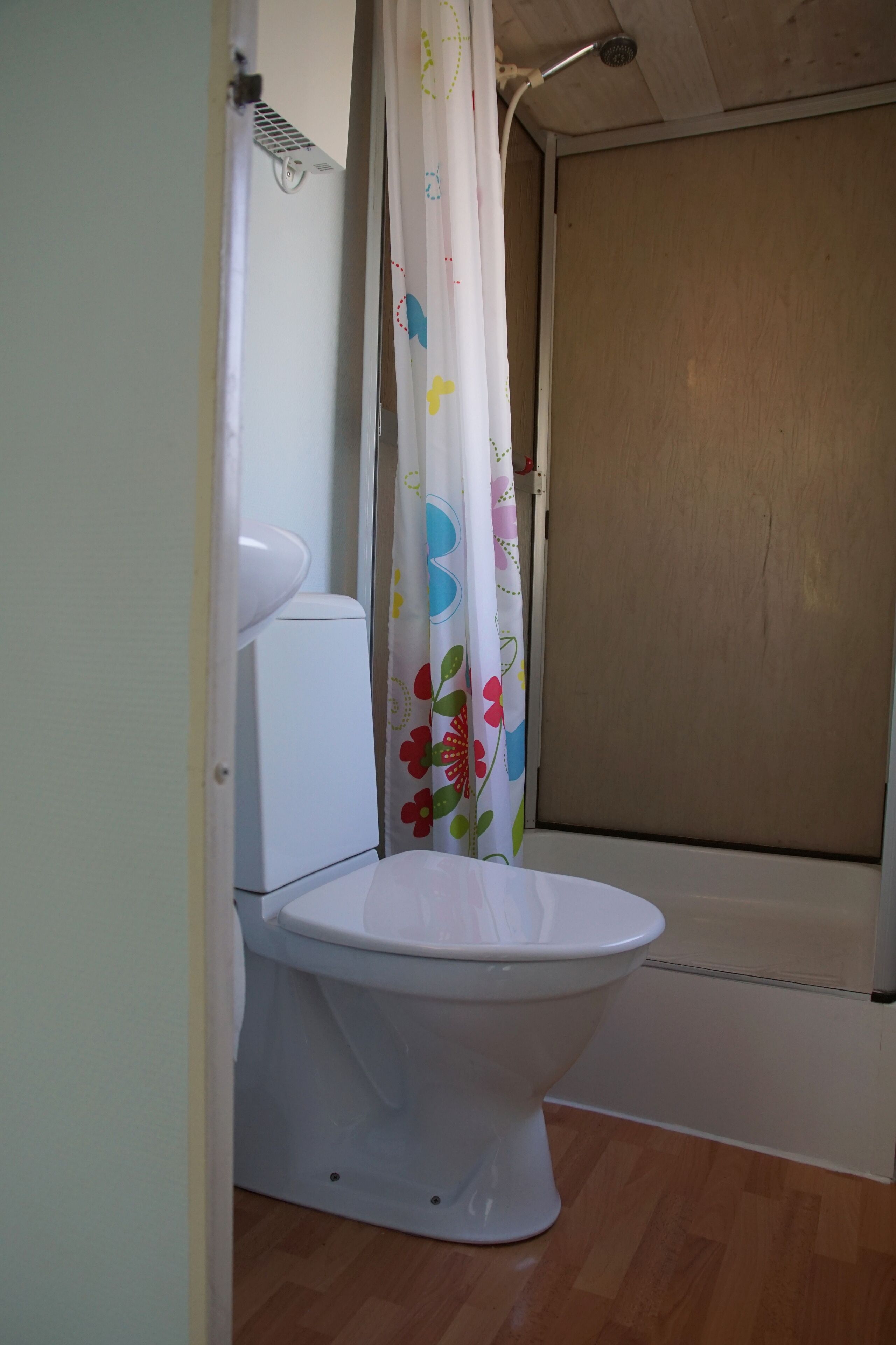 Kylpyhuone, jossa on valkoinen wc-istuin, kukkakuviollinen suihkuverho ja puulattia.