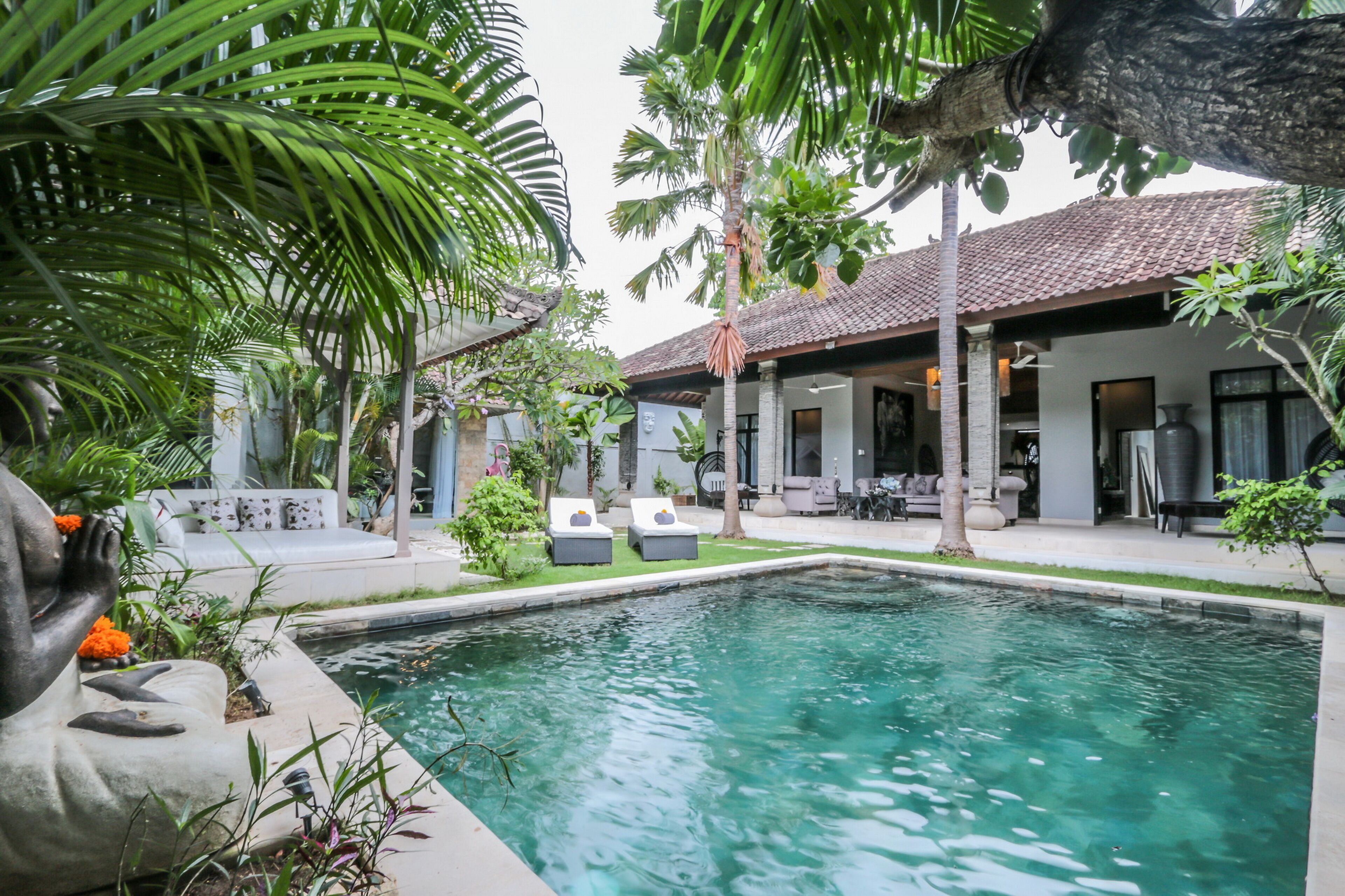 A pool area with lounge chairs, a statue, and a house with a tiled roof.
