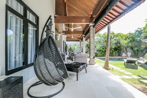 Un espace extérieur aménagé, abrité et doté de sièges confortables, d’une chaise suspendue et d’une piscine en arrière-plan.