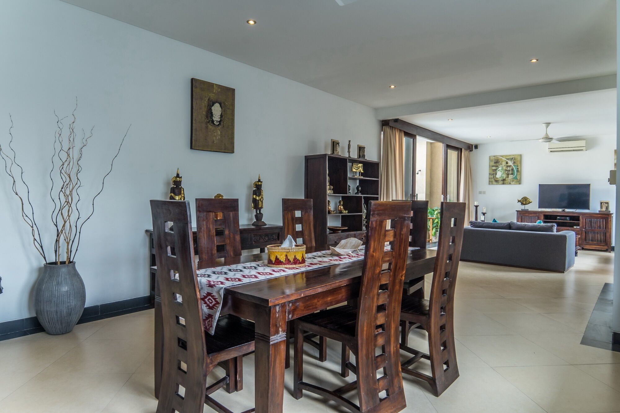 A dining area with a wooden table and chairs, a vase with branches, and a bookshelf in the background.