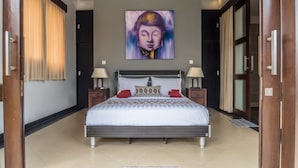 A bedroom with a large bed, two bedside tables with lamps, a painting on the wall, and a view through an open door. - Villa Kiaja (Kerobokan)