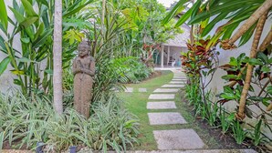Um caminho no jardim com uma estátua, cercado por diversas plantas e um edifício ao fundo.