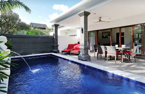 Ein Schwimmbecken mit einer roten Liege, ein Essbereich mit Rattanmöbeln und ein modernes Haus mit großen Fenstern.
