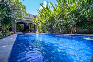 A swimming pool surrounded by lush greenery, with a covered area and a modern building in the background. - Villa Cantik (Legian)