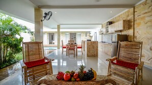 A spacious living area with two wooden chairs, a dining table with fruit, and a view of a kitchen and pool area. - Villa Airin (Canggu)