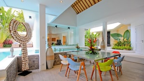 Un espace repas moderne avec une table en verre et des chaises colorées, agrémenté de plantes d’intérieur luxuriantes et offrant une vue sur une piscine.