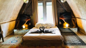 A cozy, rustic bedroom with a bed centered on a patterned rug, flanked by two lamps and a window with curtains. - Villa Wonderland (Seminyak)