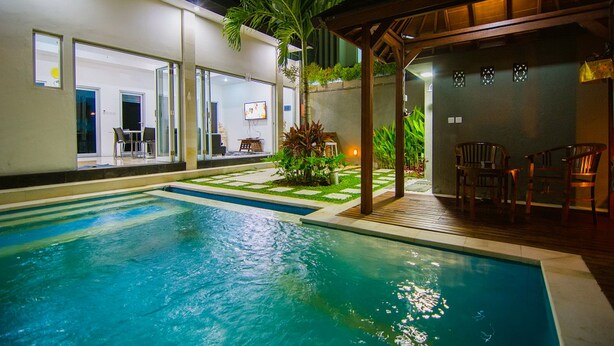 Une piscine dotée d’une terrasse en bois et de chaises, entourée d’un espace extérieur bien éclairé, avec un bâtiment visible au loin.