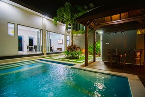 A swimming pool with a wooden deck and chairs, surrounded by a well-lit outdoor area with a building in the background. - Villa Kelapa View (Kuta)