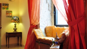 A cozy room with a window covered by red curtains, a small table with a lamp, a suitcase, and a decorative item. - Chantale's posada (Giovinazzo)