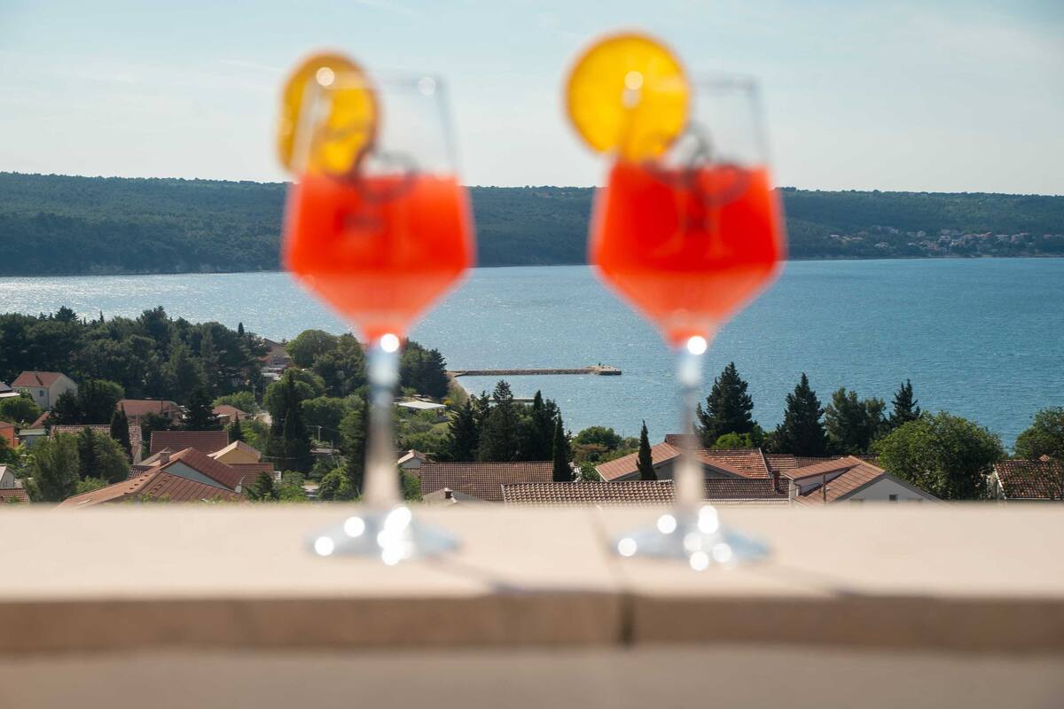 Deux verres d’une boisson rouge, garnis de rondelles de citron, sont posés sur un balcon donnant sur un lac et des maisons.