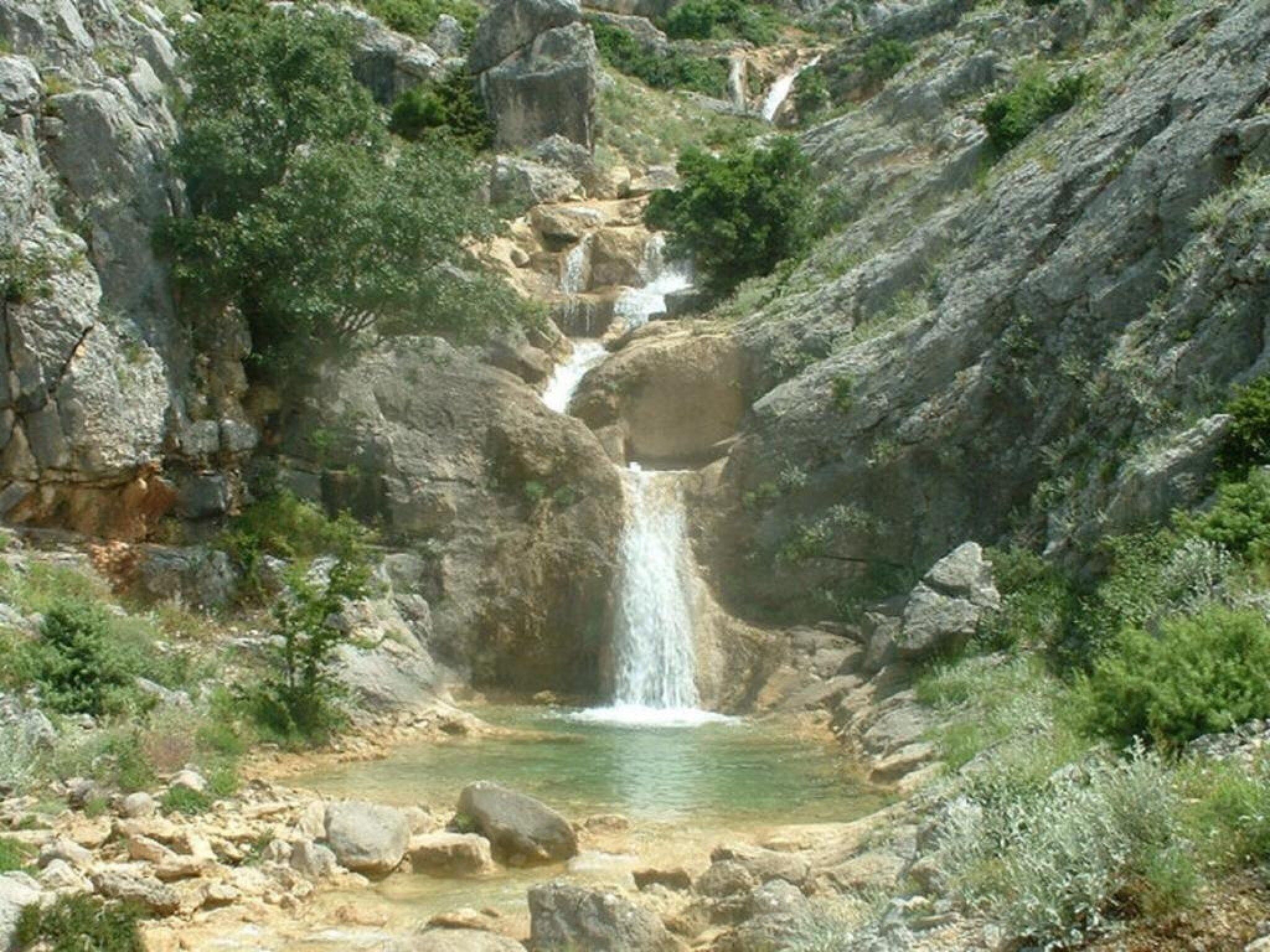 A natural waterfall cascading into a rocky pool surrounded by rugged terrain and sparse vegetation.