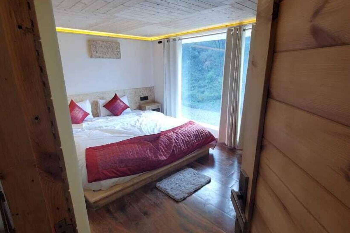 Ein Schlafzimmer mit Holzbalkendecke, einem Bett mit roter Bettwäsche, einem Nachttisch und einem Fenster mit Vorhängen.