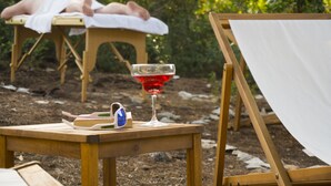 Una mesa de madera con un vaso de bebida roja, un par de gafas de sol y un libro. Al fondo, una persona recostada en una mesa de masaje al aire libre.