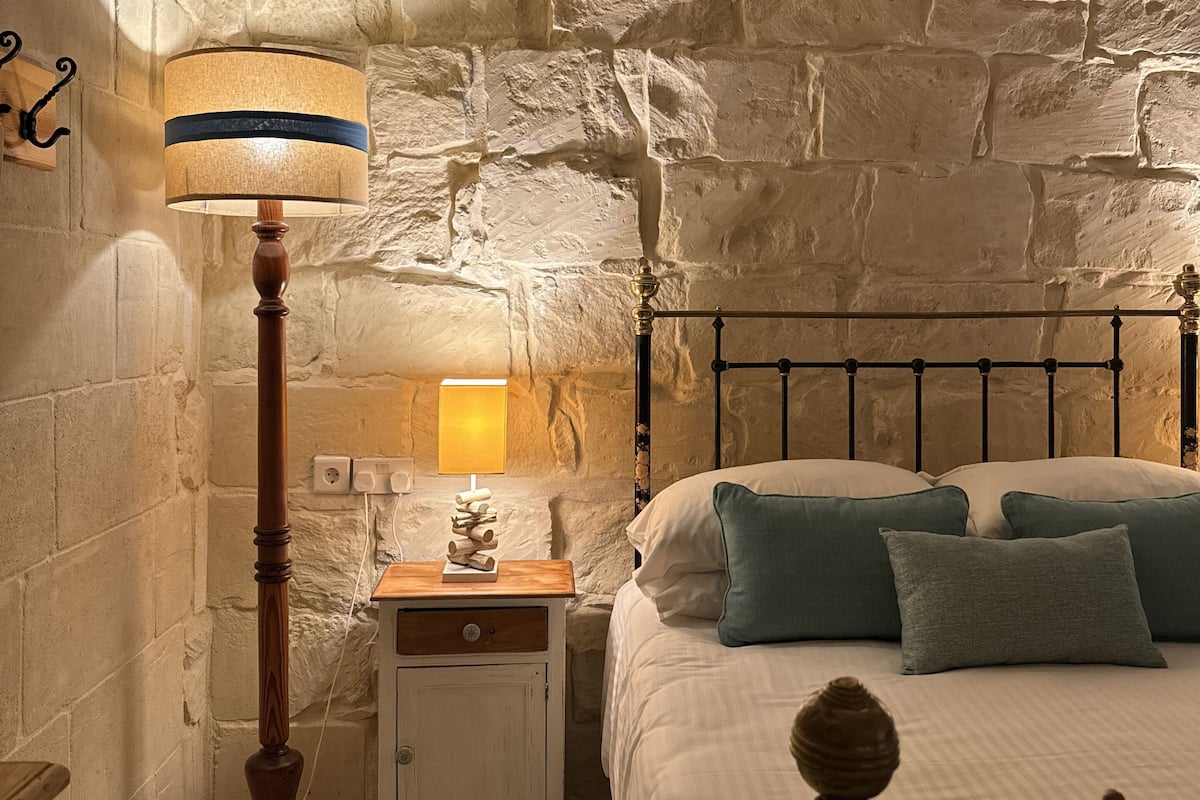 A bedroom with a stone wall, a wooden floor, a bed with pillows, a bedside table with a lamp, and a nightstand with a decorative item.
