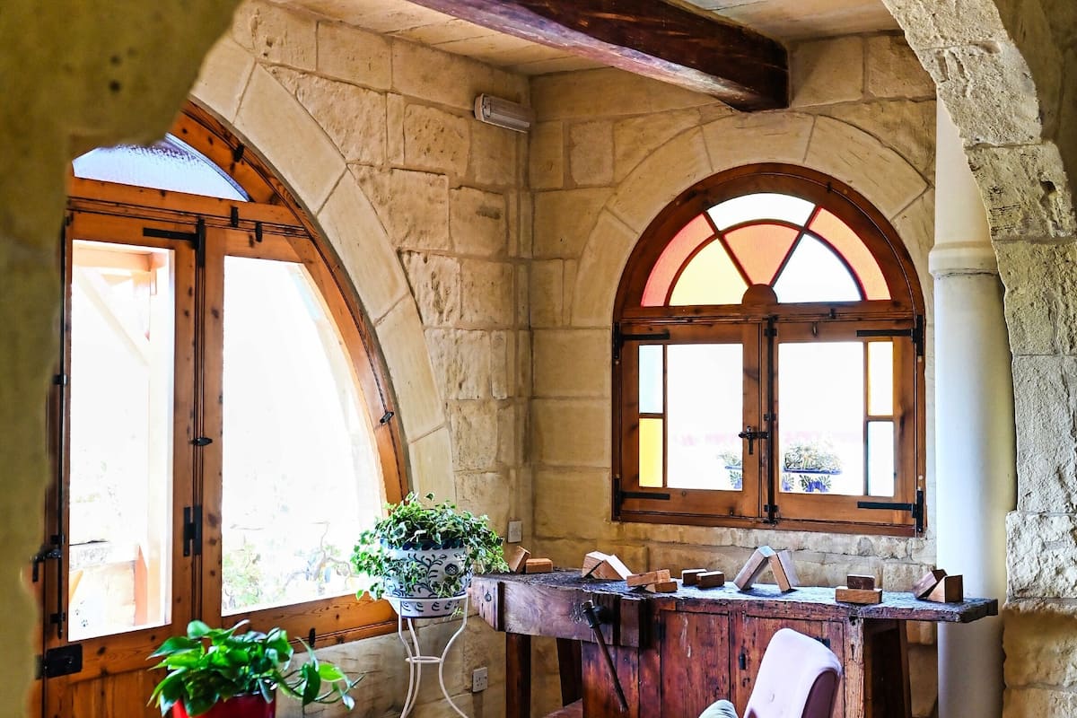 A room with stone walls, a wooden table, a chair, and a window with an arched design.