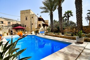 A swimming pool with a stone building in the background, palm trees, and outdoor seating. - Maria's B&B (Ghasri)
