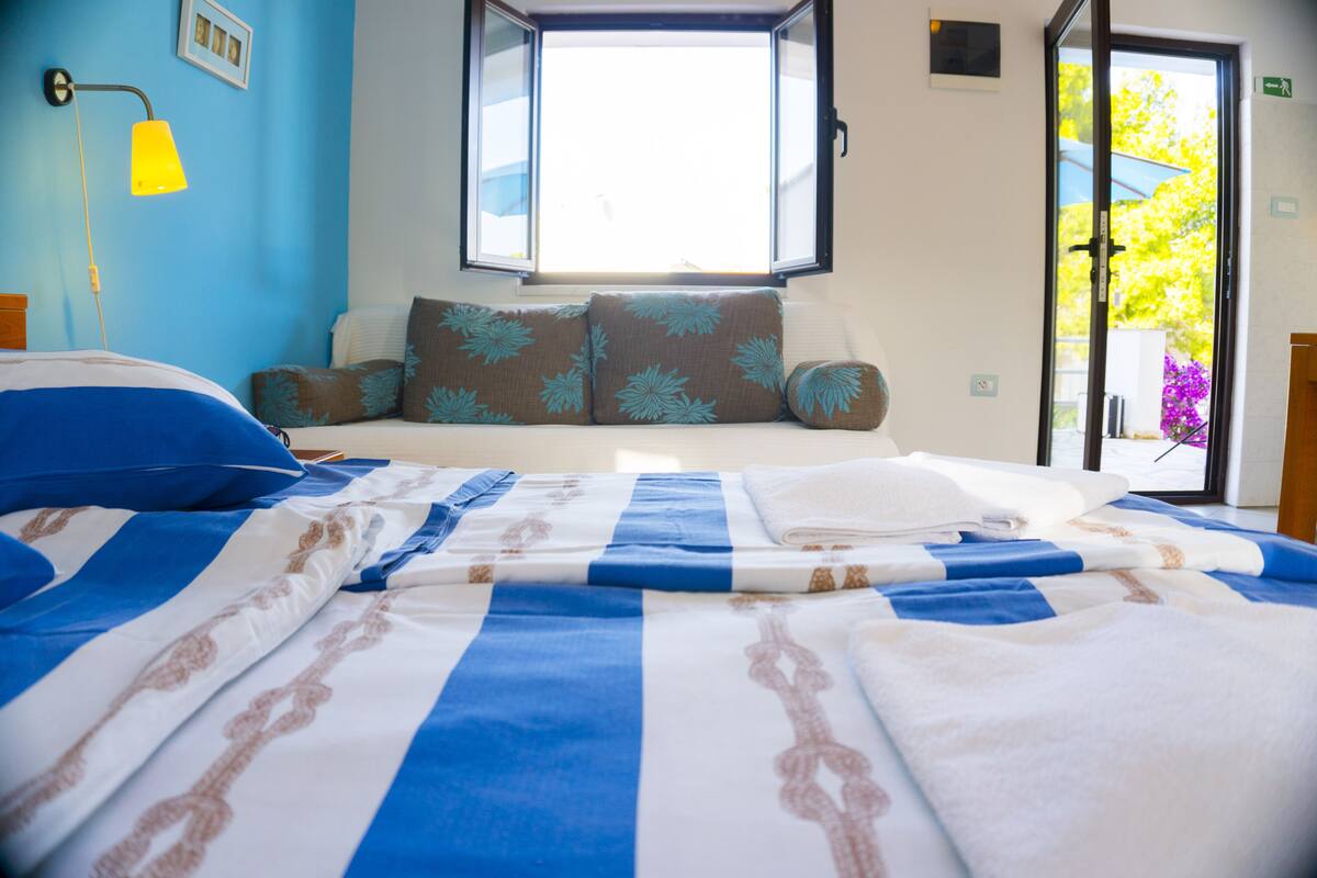 A neatly made bed with blue and white bedding, a window with a view of greenery, and a yellow wall lamp.