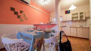 Un comedor con una mesa dispuesta para dos, sillas blancas, y una cocina con armarios y una encimera a la vista en el fondo.