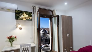Ein Schlafzimmer mit einem Bett, einem Schreibtisch mit Stuhl, einem Kleiderschrank und einem Balkon mit Blick auf Gebäude.