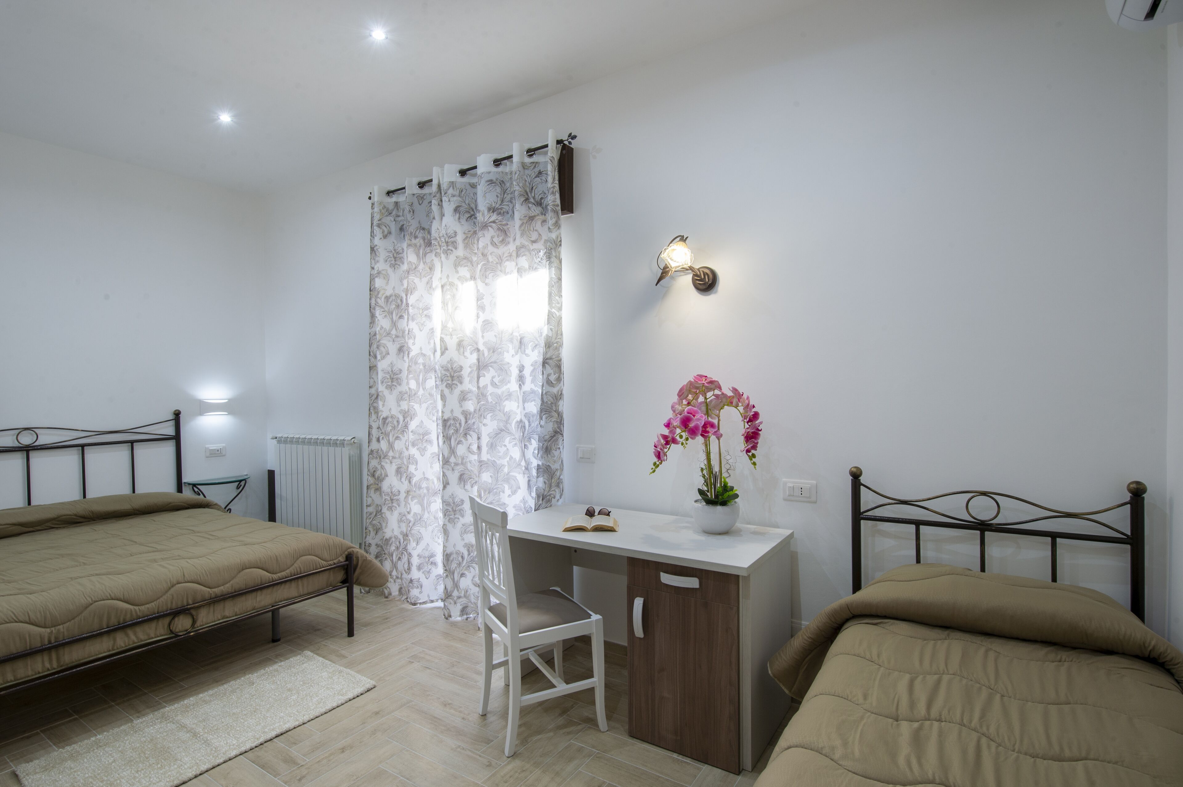A bedroom with two beds, a desk, a chair, a radiator, and a curtain.
