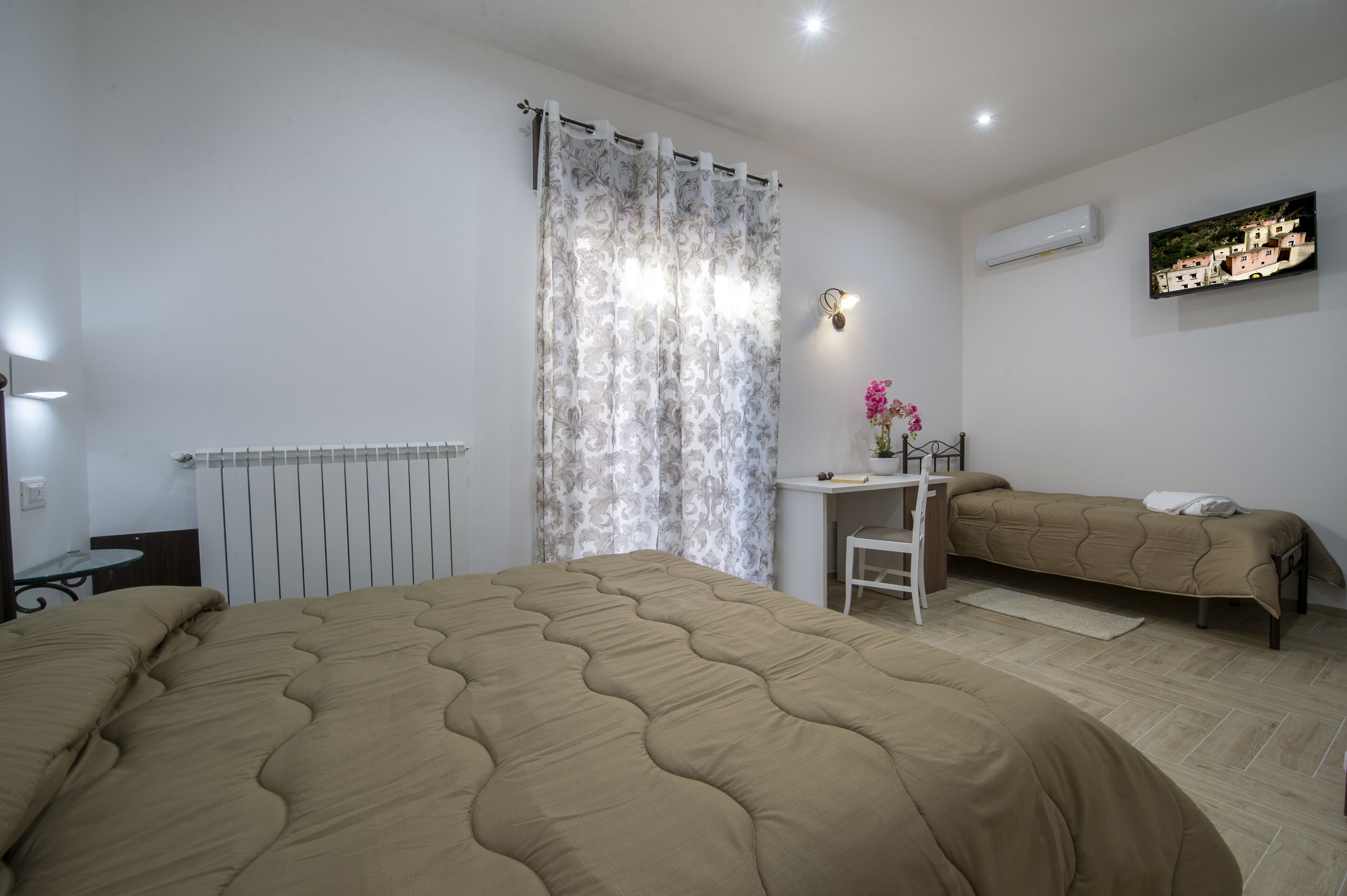 Een slaapkamer met een groot bed, een bureau met stoel, een klein nachtkastje, een radiator, een gordijn en een wandairconditioner.