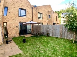 Una casa de ladrillo con patio trasero cercado, un patio con mesa y sillas, y un pequeño jardín.