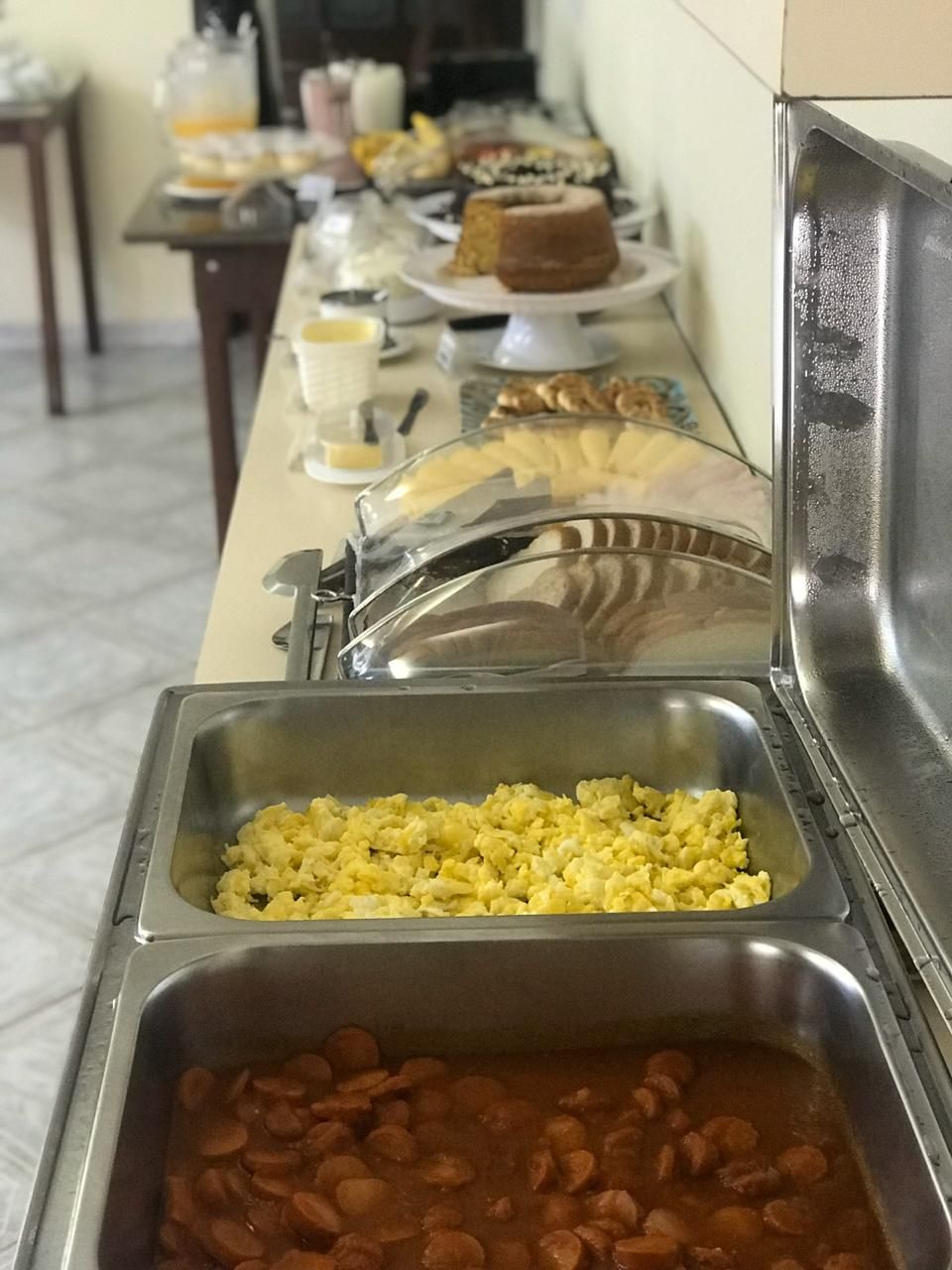 Montaje de bufé con diversas opciones de comida, entre ellas huevos revueltos, salchichas y un pastel.