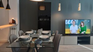Un espace repas moderne avec une table en verre pour quatre personnes, un téléviseur diffusant une émission avec deux personnes et des luminaires suspendus.