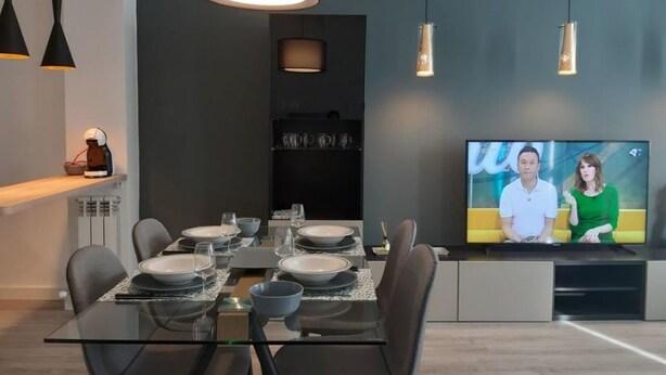 Un comedor moderno con una mesa de vidrio preparada para cuatro personas, un televisor que muestra a dos personas y luminarias colgantes.
