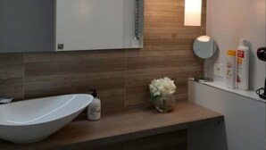 Un baño moderno con un espejo grande, paneles de madera en la pared, un lavabo blanco, un espejo redondo y un florero con flores blancas.