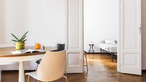 A room with a wooden dining table, a chair, a potted plant, and a bed with a white headboard. - Macchi Master Guest apartment (Milan)