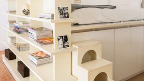 A modern, multi-tiered bookshelf with books and framed photos, positioned next to a bed in a minimalist room. - Siri Master Guest apartment (Milan)
