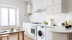 A modern kitchen with white cabinetry, a built-in oven, a washing machine, and a window with blinds. - Siri Master Guest apartment (Milan)