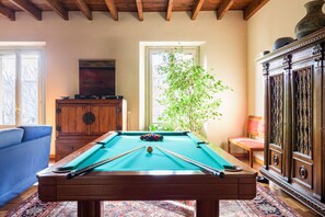 A pool table in a room with a wooden ceiling, a cabinet, a chair, and a large window with a view of trees.