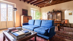 A living room with a blue sofa, wooden coffee table with books, a dining table with chairs, and a large window with a view of buildings. - Morgagni Master Guest apartment (Milan)