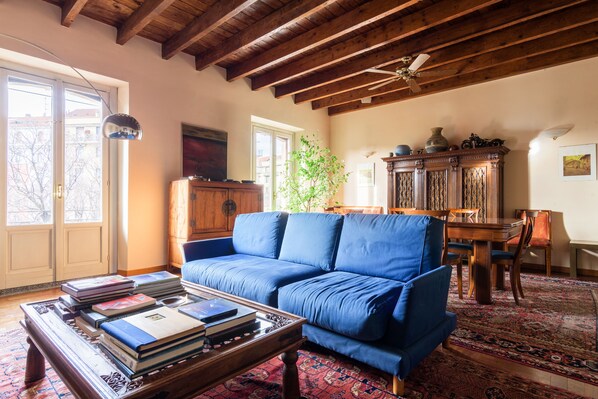 A living room with a blue sofa, wooden coffee table with books, a dining table with chairs, and a large window with a view of buildings.