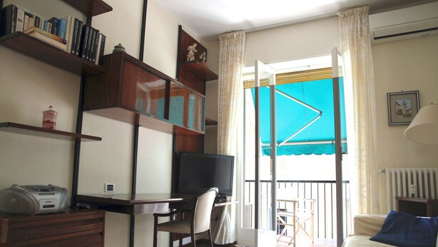 Ein Wohnzimmer mit Fernseher, Schreibtisch und Sessel. Regale mit Büchern und einem roten Behälter sind vorhanden. Ein Balkon mit blauer Markise ist sichtbar.