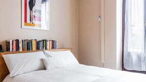 A neatly made bed with white linens, a bookshelf with various books, and a poster on the wall. - Asso Master Guest apartment (Milan)
